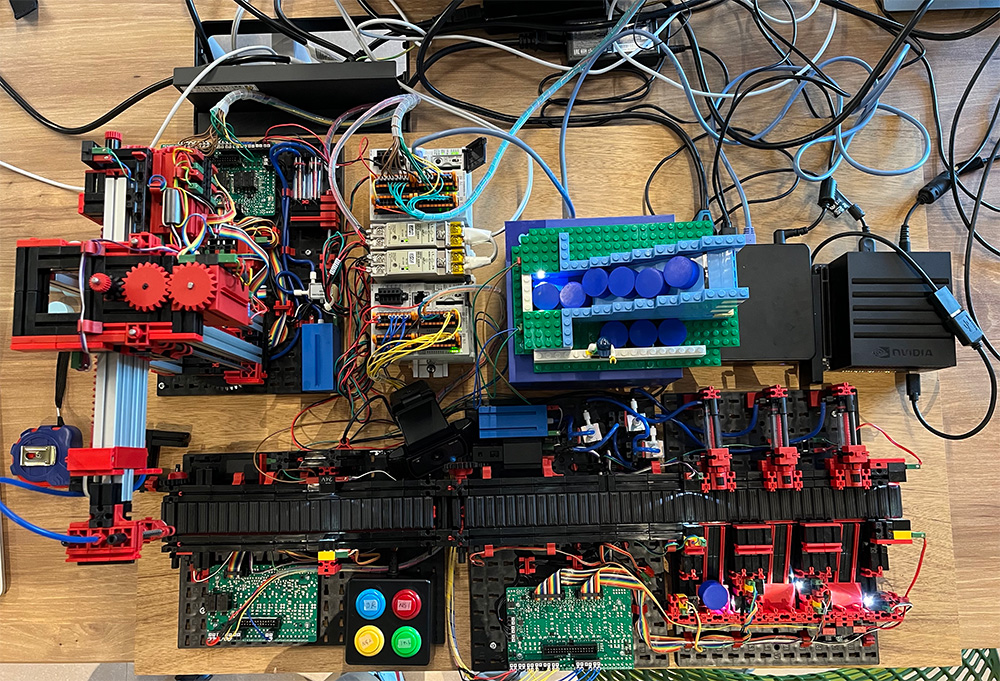 Top-down view of a digital twin factory model featuring various electronic components, wiring, control boards, colorful gears, and push buttons, set up on a wooden surface. The setup includes red, blue, black, and green plastic parts and numerous connecting cables, illustrating an advanced industrial automation system.