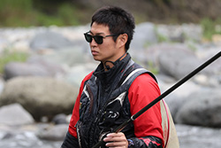 A person wearing sunglasses and outdoor gear is fishing by a rocky riverbank.