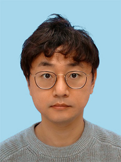 A portrait of a person with short brown hair, wearing round glasses and a gray sweater, against a light blue background.