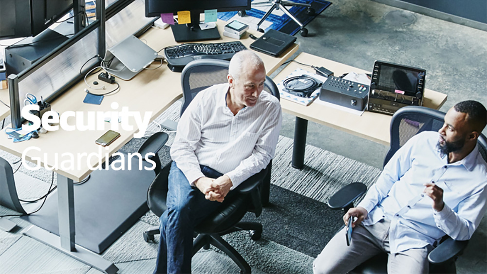 Two professionals having a discussion in a modern office environment with multiple computer monitors and office equipment, representing the concept of security guardians.