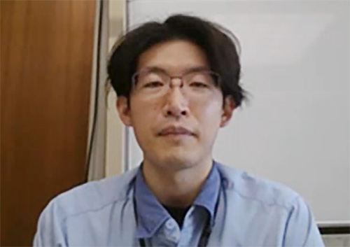 Portrait photo of a man with glasses and dark hair, wearing a light blue shirt, seated indoors in front of a whiteboard and wooden panel.