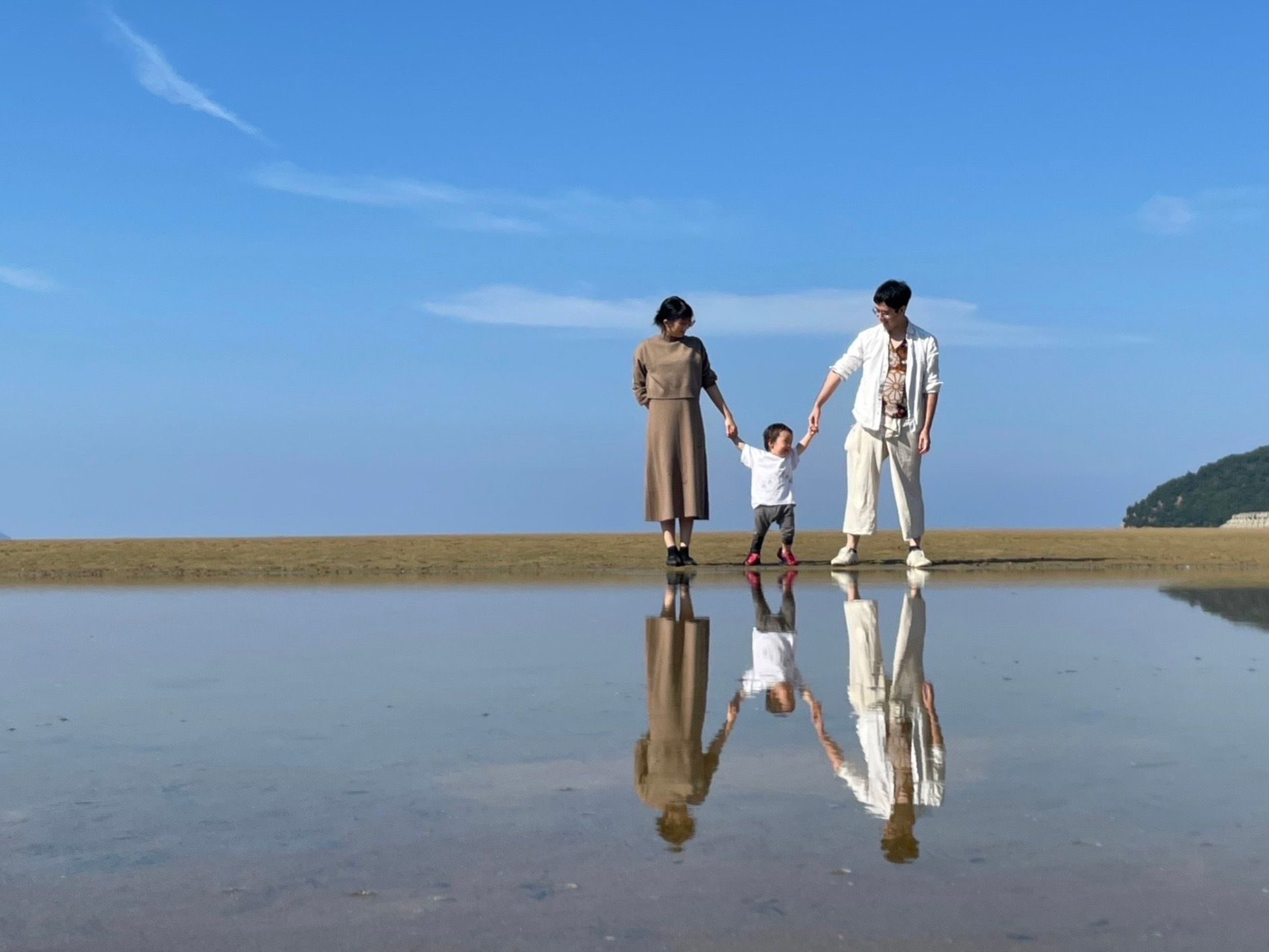A family holding hands, walking on a sandy beach with a clear reflection of themselves in a shallow pool of water under a bright blue sky.