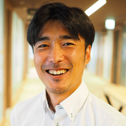 A man smiling in an office setting, wearing a white shirt. The background shows a modern office hallway with natural light.