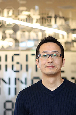 A portrait photo of an individual wearing glasses and a dark sweater, standing in front of a blurred office background with abstract patterns and glass walls.