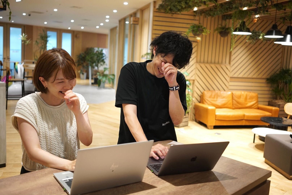 Two young adults laugh and enjoy working together on their laptops in a modern coworking space with plants, wooden decor, and a comfortable lounge area.