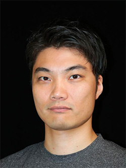 Portrait photo of a man with short black hair wearing a gray shirt, set against a black background.