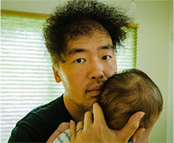 A man holding a baby close to his shoulder, looking directly at the camera in a softly lit room.