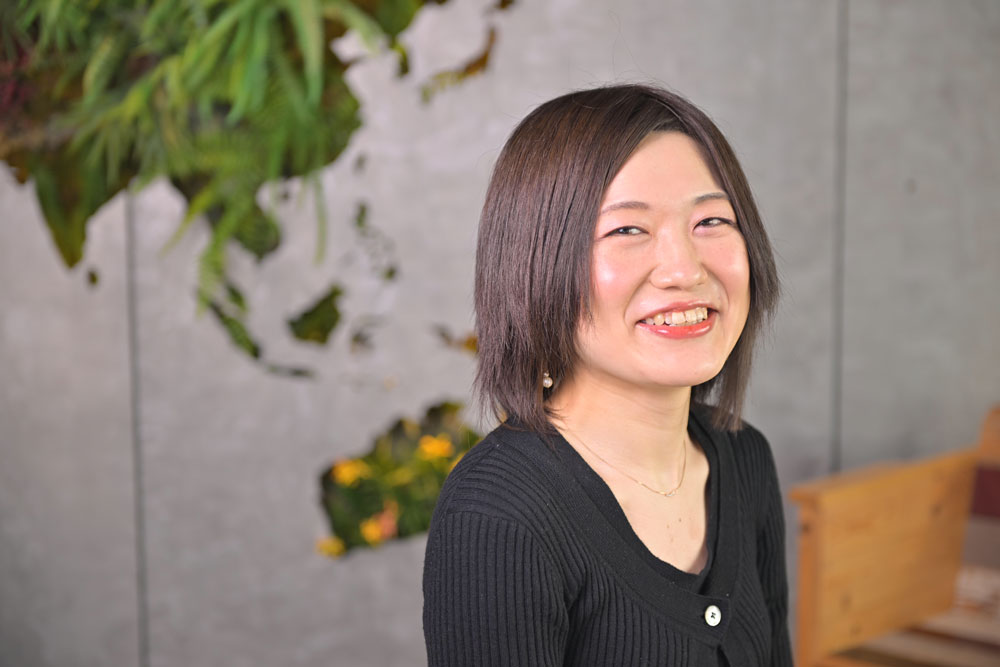 A woman smiling indoors with a modern background featuring plants and a wall design.