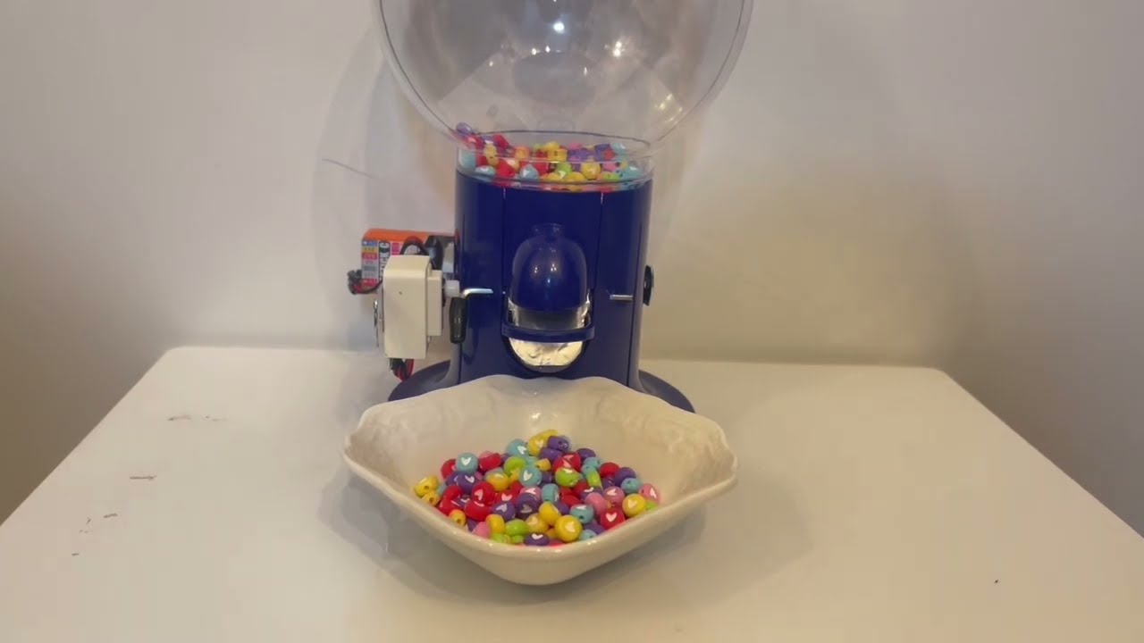 A blue candy dispenser machine dispensing colorful heart-shaped candies into a white bowl, placed on a white surface with a plain background.