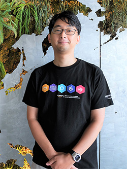 A person wearing glasses and a black AWS t-shirt with colorful icons is standing in front of a wall with greenery and a map pattern. The setting appears casual and modern.