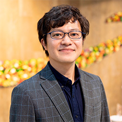 A portrait of a man wearing glasses and a plaid blazer, standing in an indoor setting with a blurred background.