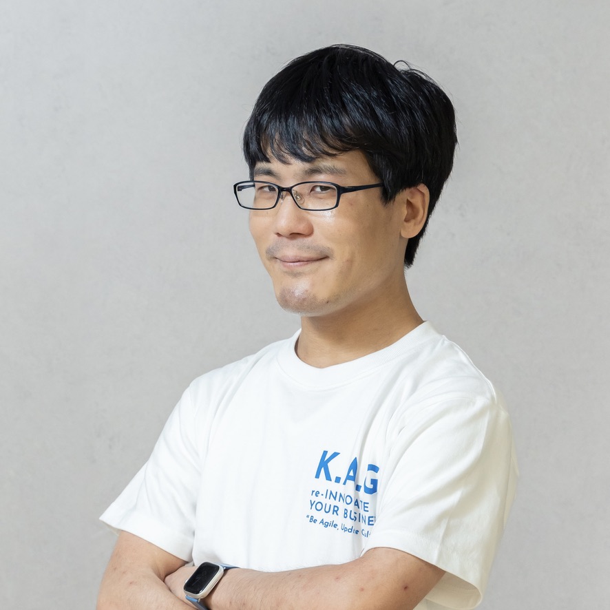 A person wearing glasses and a white t-shirt, crossing their arms and smiling, standing against a plain background.