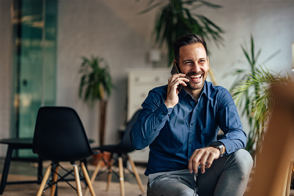 A man in a blue shirt is smiling while talking on a smartphone, seated in a modern office environment with plants and contemporary furniture in the background.