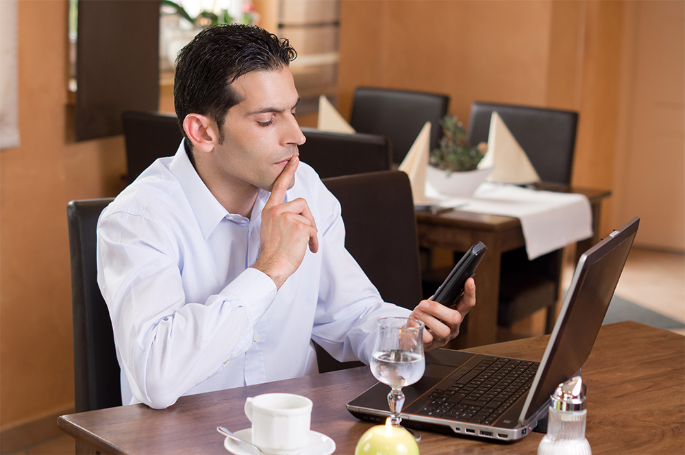 A man sits at a table in a cafe, thoughtfully looking at his smartphone while using a laptop, with a cup of coffee and a glass of water in front of him.