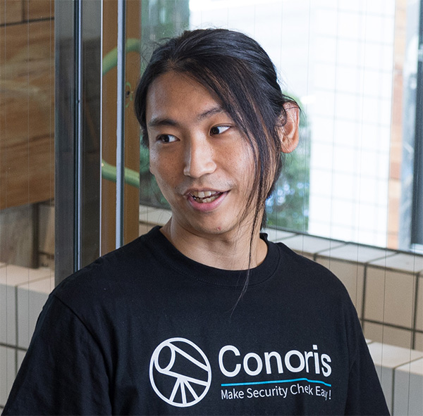 A person with long hair tied back is wearing a black 'Conoris' t-shirt and standing in front of a glass wall with tiled patterns.