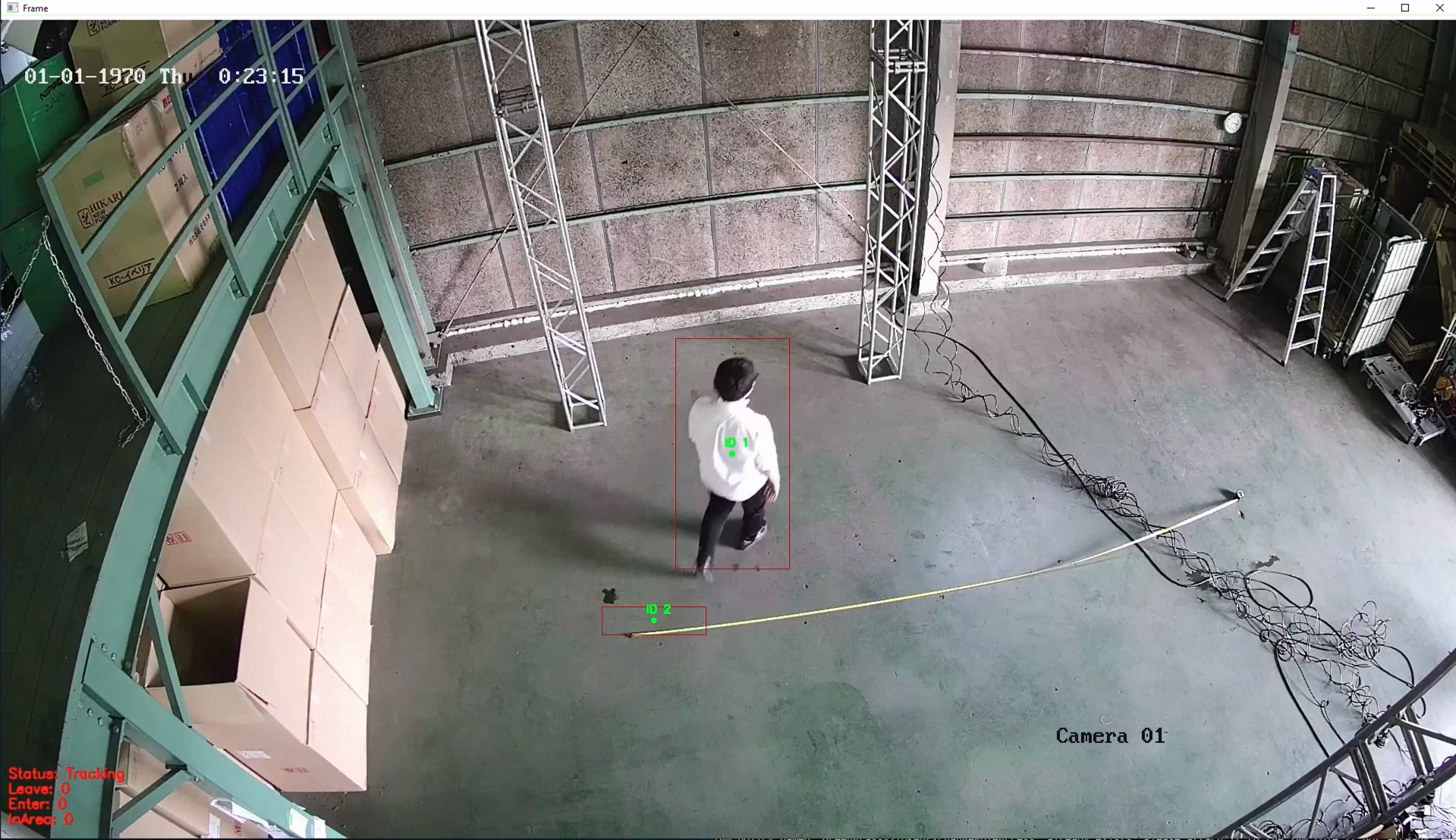 A security camera image from a warehouse shows a real-time tracking system in use. A person is walking across the floor, identified by a tracking ID and highlighted by a box on the screen. The system displays statistics for tracking, entrances, and exits in red text. Large cardboard boxes are stacked on one side, and various warehouse equipment can be seen.