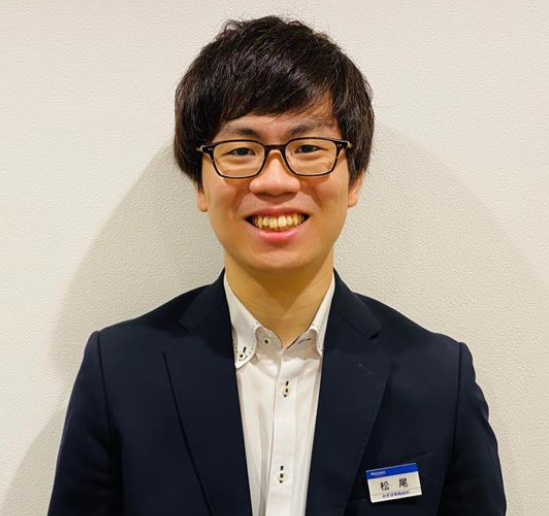 A person wearing glasses and a suit jacket, smiling while standing against a plain light-colored wall.