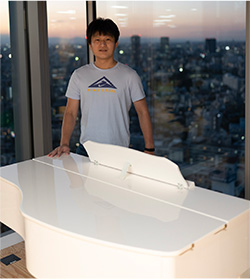A person is standing behind a white grand piano in front of large windows, with a cityscape visible at sunset in the background.