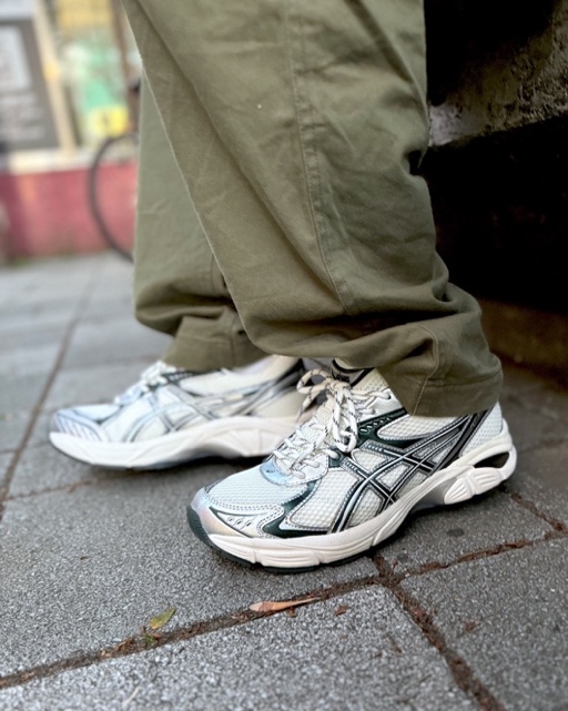 A close-up image of a person wearing green pants and white running shoes, standing on a city sidewalk.