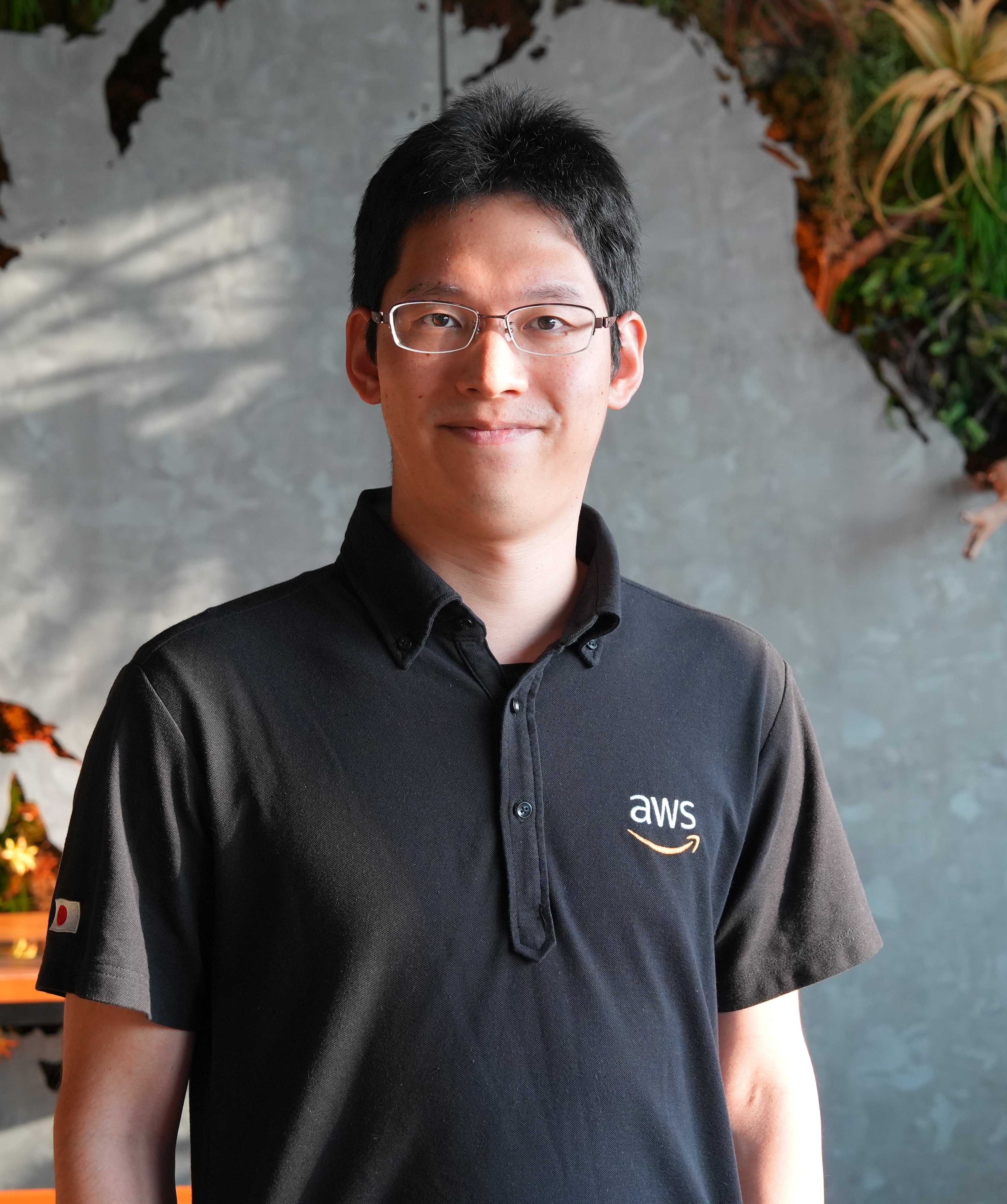 Portrait of an AWS employee in the Japan office, wearing an AWS-branded shirt and standing in front of a decorative wall with plants.