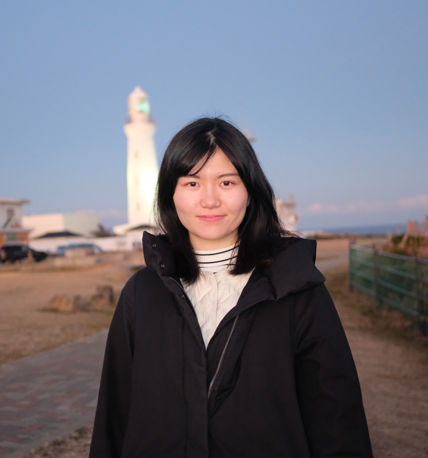 A woman wearing a black coat stands outdoors near a lighthouse at dusk, with the sky transitioning to evening.