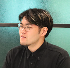 A man wearing glasses and a black shirt, sitting in front of a green background.