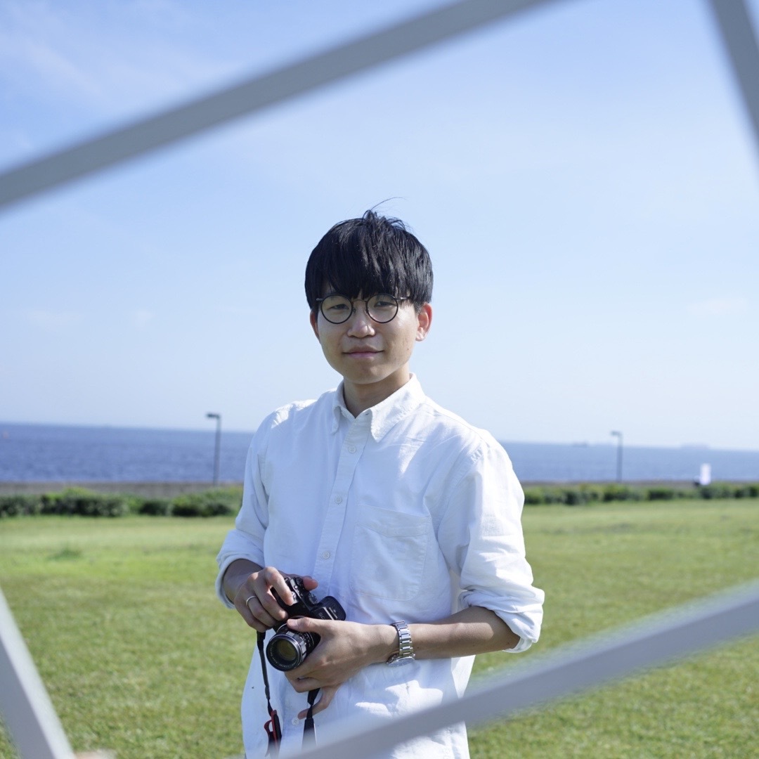 A person in a white shirt holding a camera while standing on grass in a seaside park, with the ocean and blue sky in the background.