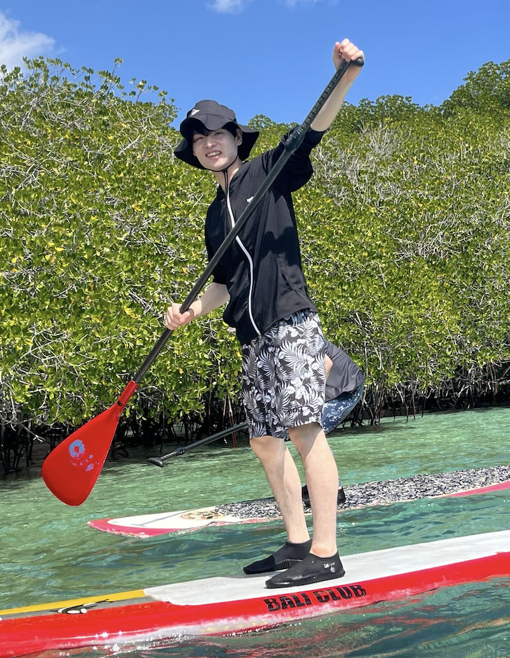 A person paddleboarding on clear tropical water near lush mangroves under a blue sky.
