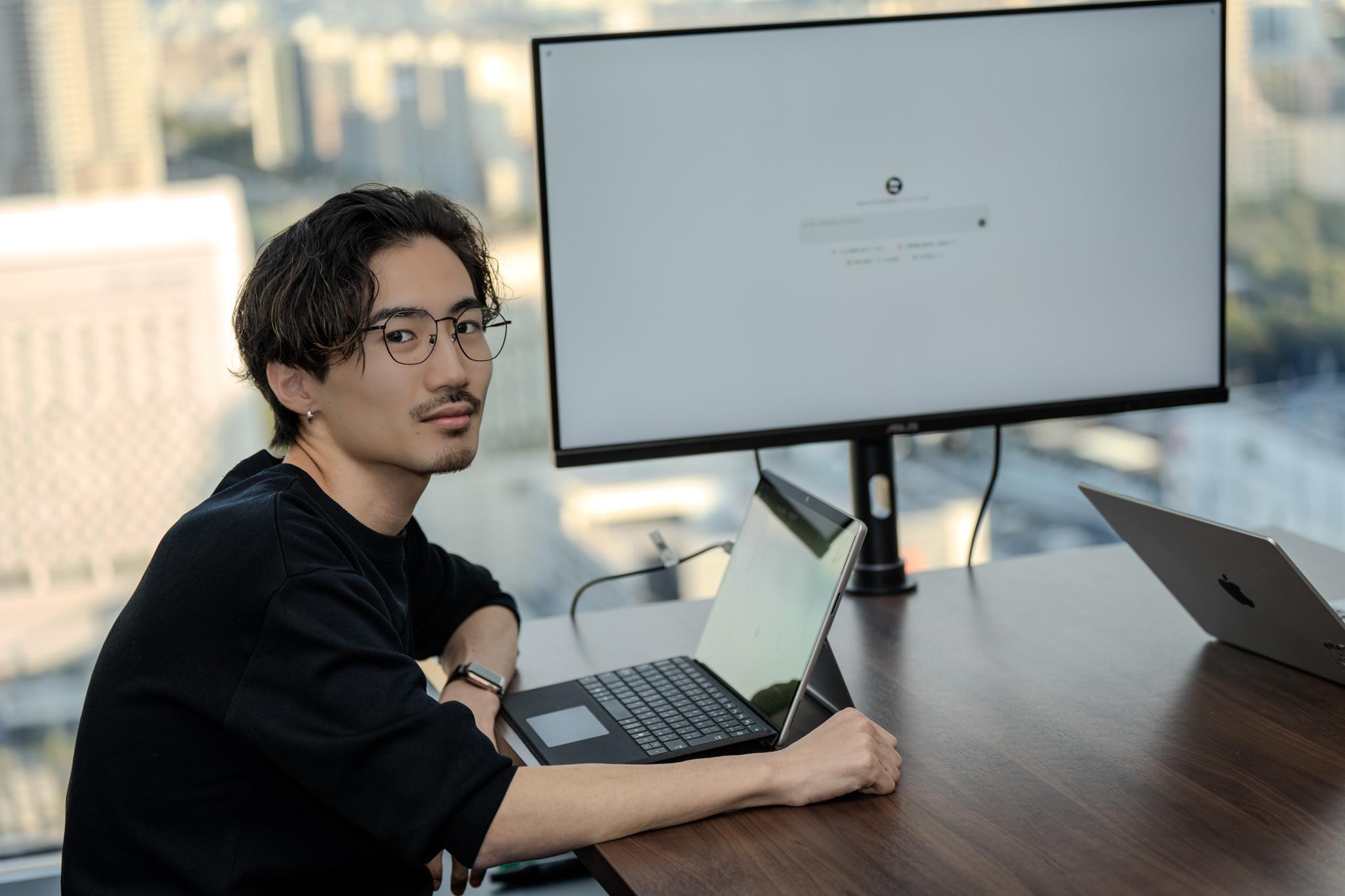 A man is sitting at a desk in a modern office, working on a laptop with a large monitor in the background. The setting includes city buildings seen through the window.