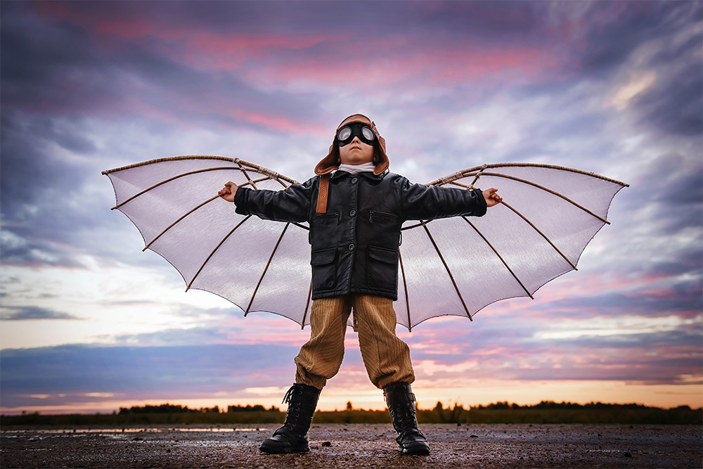 A child dressed in an aviator costume with DIY wings stands with arms outstretched against a colorful sunset sky, embodying imagination and the spirit of adventure.