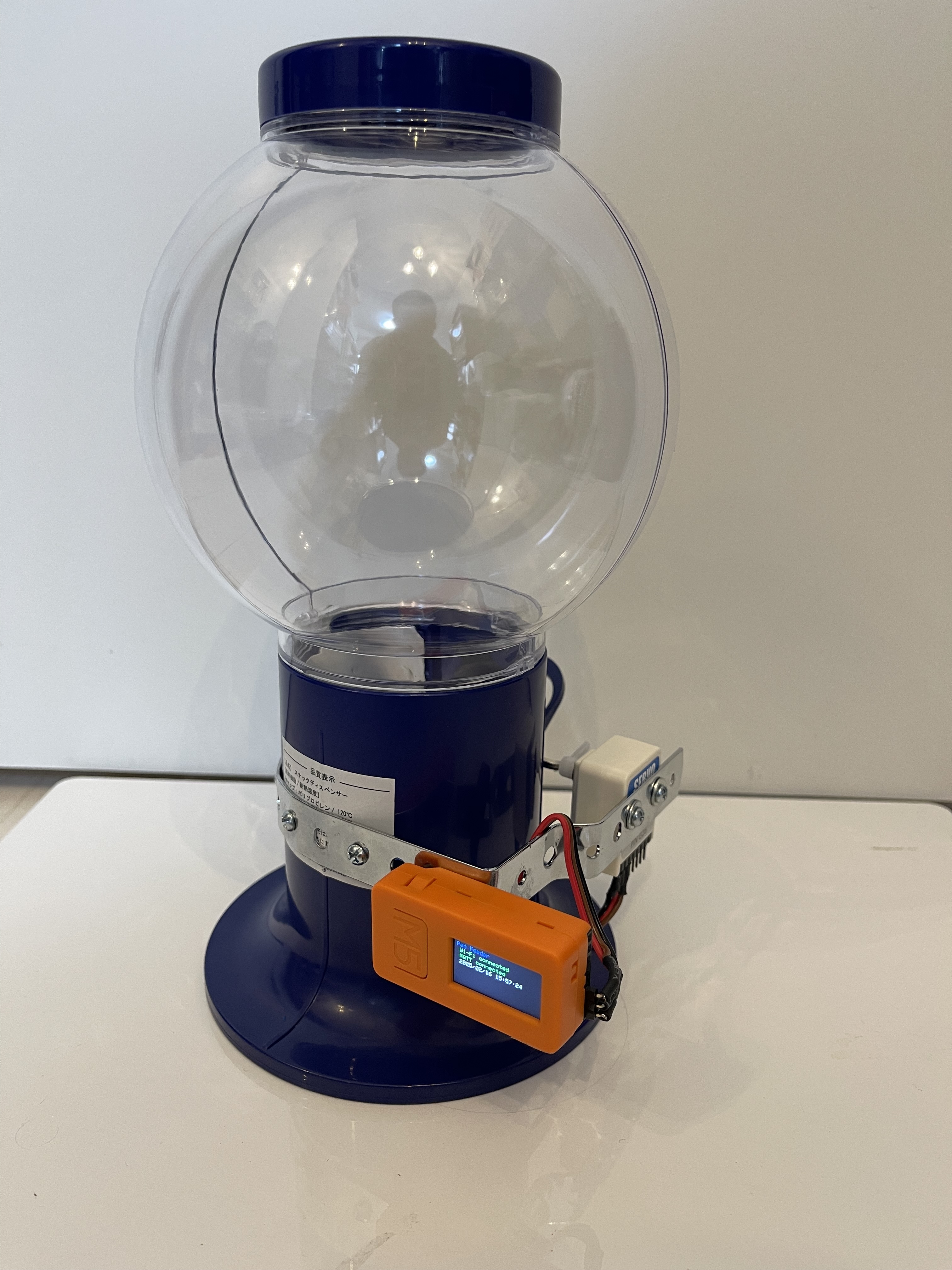 A transparent round gumball or candy machine with a blue base and lid, featuring an attached orange electronic device and wiring, possibly used for automated counting or dispensing.