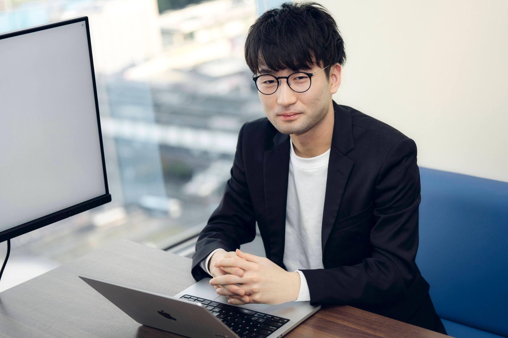 A businessman wearing glasses and a blazer sits at a desk with a laptop in a modern office setting with large windows.