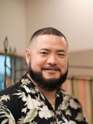 A man with a beard and short hair, wearing a black floral shirt, is smiling and looking at the camera indoors.