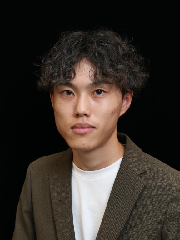 A portrait of a young man with curly hair wearing a blazer, photographed against a black background.