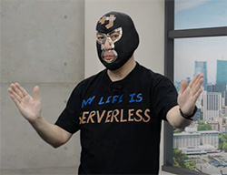 A person wearing a black wrestling mask and a T-shirt that reads 'My Life is Serverless', standing indoors with arms raised, near a window with a city view.