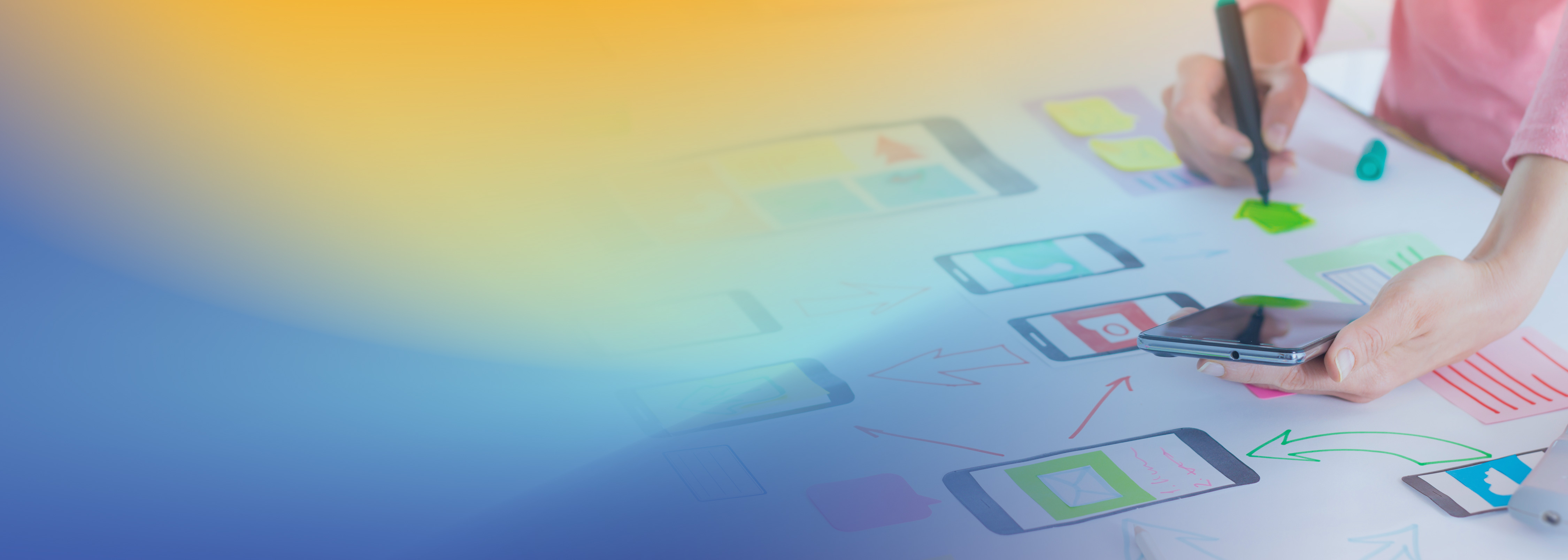 A person sketching mobile app design prototypes on paper, holding a smartphone. Various phone screen mockups and colorful sticky notes are visible, representing the process of application design and development.