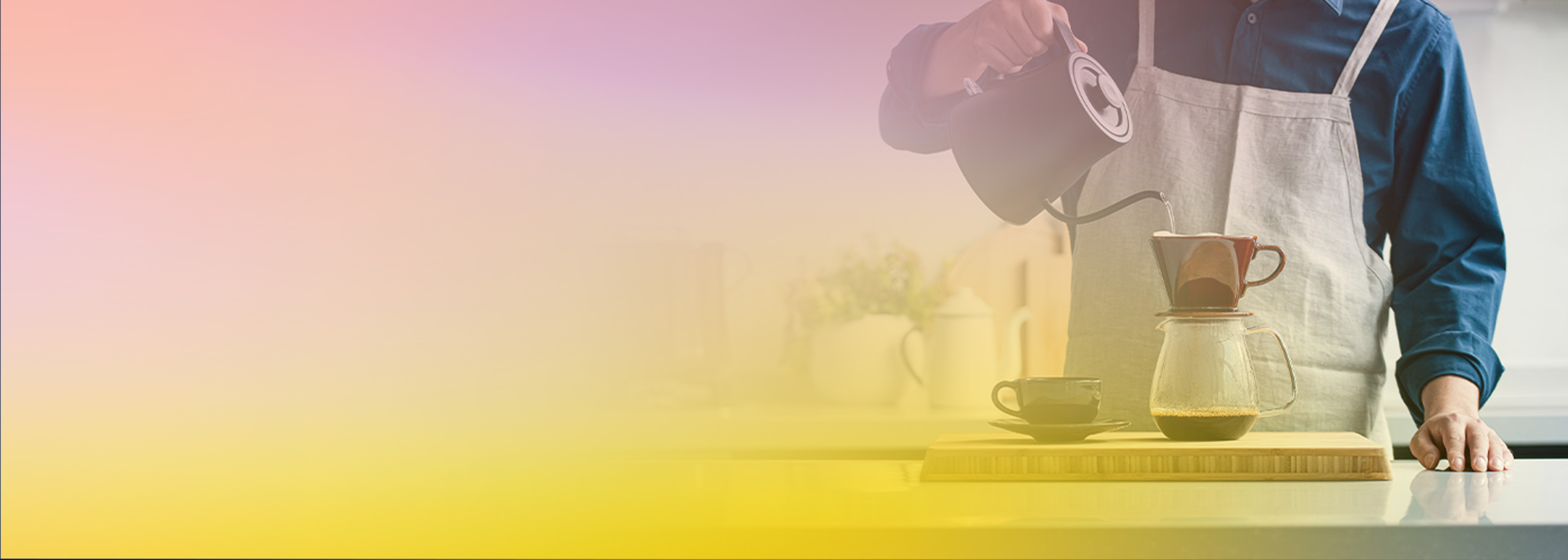 A person wearing an apron pours hot water from a kettle into a pour-over coffee maker on a counter, with soft pastel lighting and a blue shirt visible.