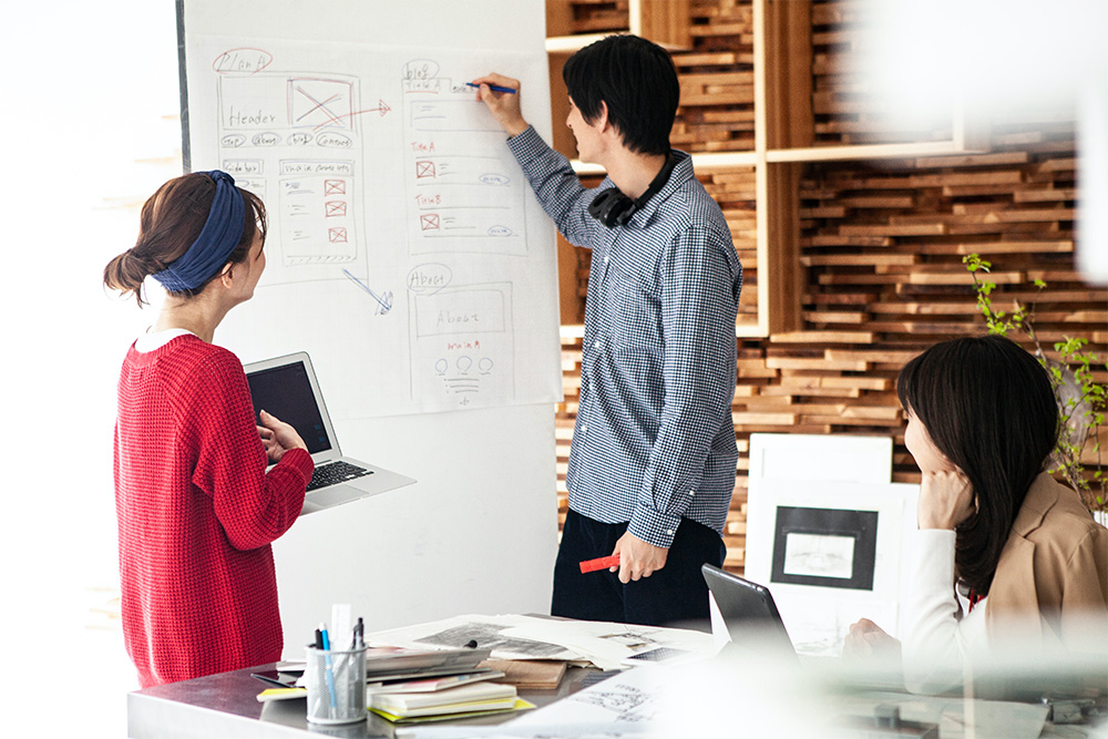 A group of people collaborating on website architecture, with sketches and wireframes on a whiteboard. One person is writing on the whiteboard while others discuss and reference laptops in a modern workspace.