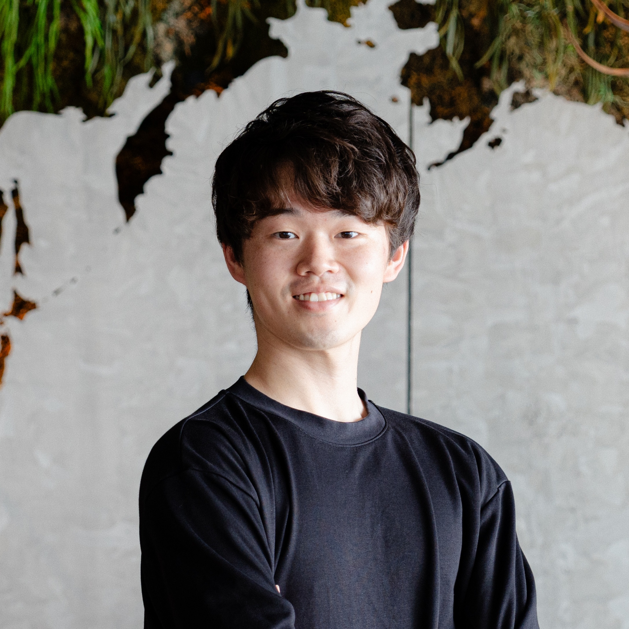 A young man wearing a black shirt standing in front of a modern, artistic background with greenery and textured wall elements.