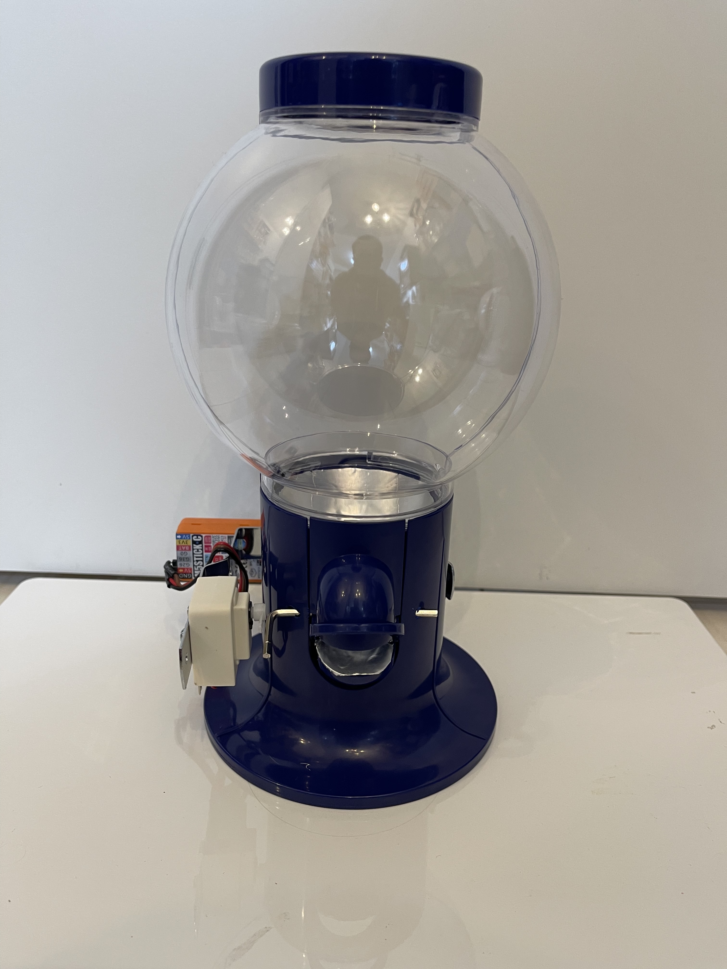 A clear plastic gumball or candy dispenser with a blue base, featuring an attached electronic component, placed on a white background.