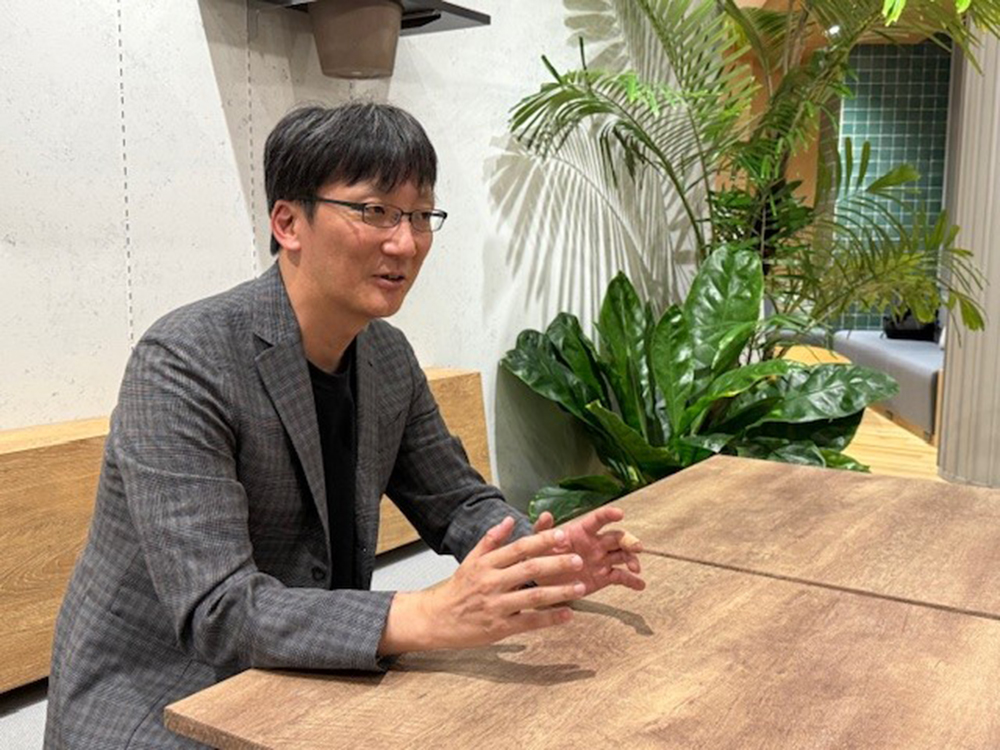 A man wearing glasses and a checked blazer is talking while seated at a wooden table, with large green indoor plants in the background.