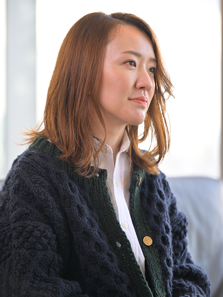 A woman with shoulder-length hair wearing a blue knit cardigan and white shirt is sitting indoors, looking thoughtful.