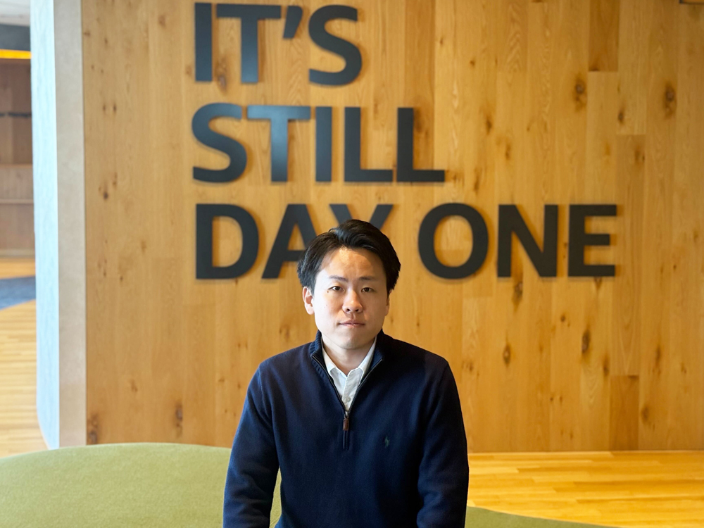 A man is sitting in front of a wooden wall with large black letters displaying the phrase 'IT'S STILL DAY ONE'.