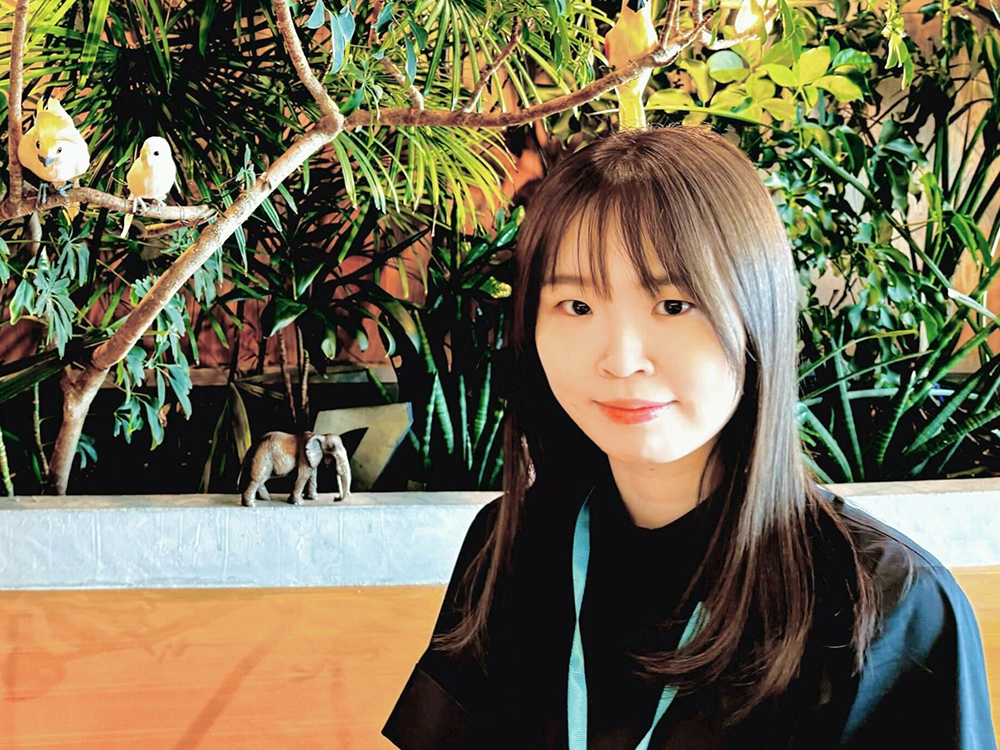 A woman sitting in a cafe surrounded by lush green plants, with small birds on branches in the background.