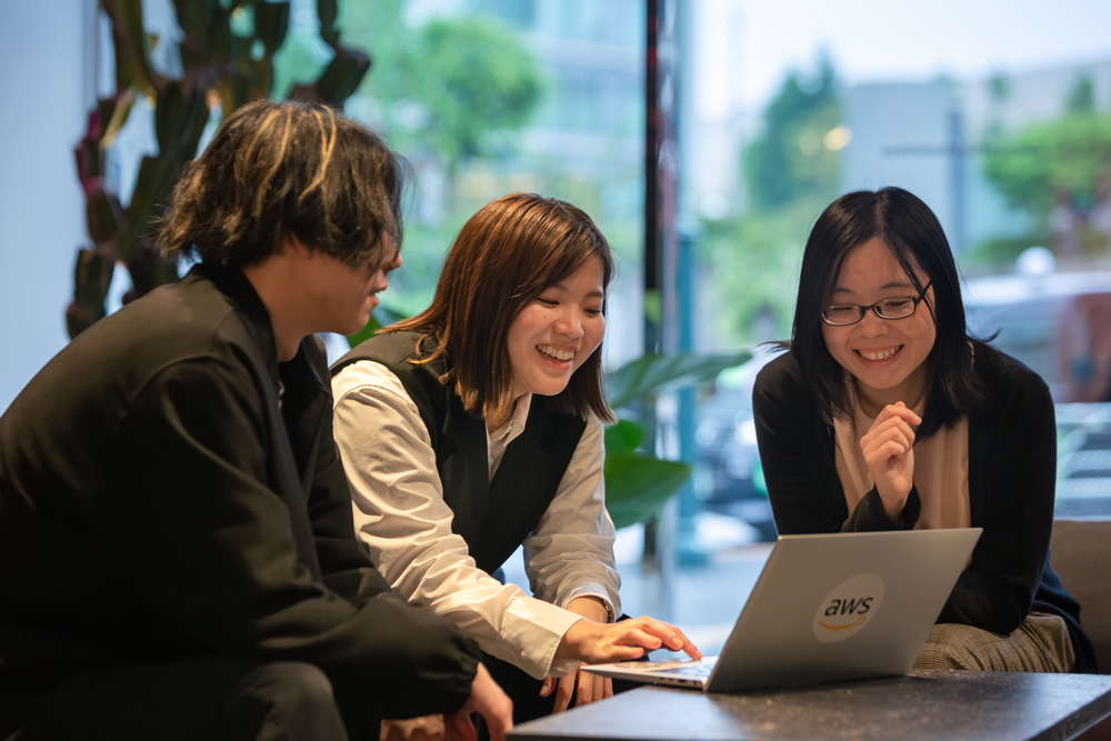Three people collaborating around a laptop with an AWS logo in a modern office setting.