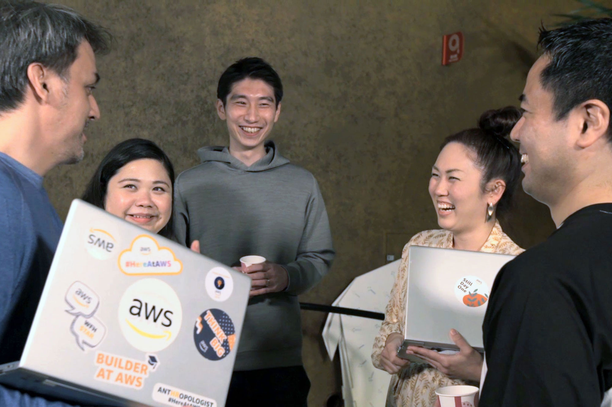 A group of AWS employees are collaborating and smiling together in a casual setting, each with laptops that have AWS-themed stickers.