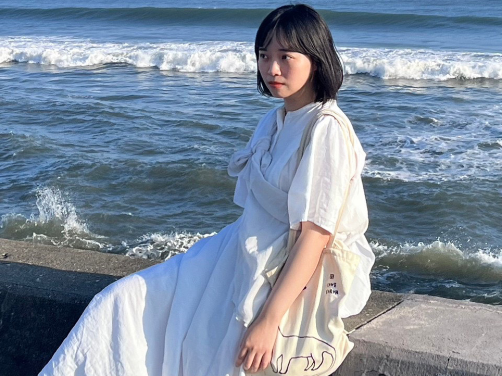 A woman in a white dress sitting on a concrete edge near the sea, with waves in the background.