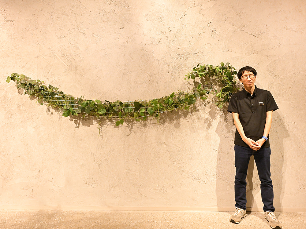 A person wearing an AWS shirt stands next to a wall decorated with a horizontal arrangement of greenery. The background is a textured beige wall, and the atmosphere appears casual and professional.