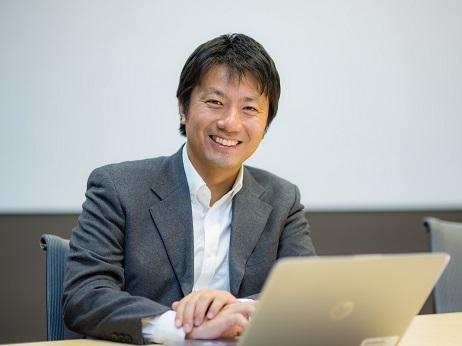 A professional man in a business suit is smiling while seated at a meeting table with a laptop in front of him.