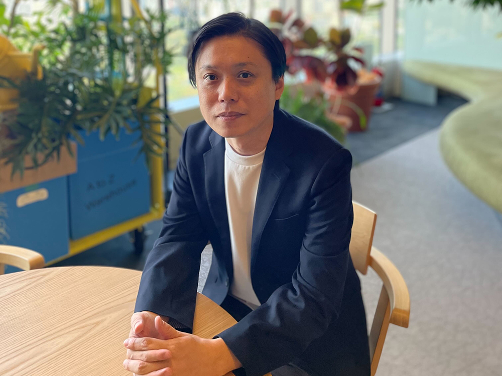 A man in a black blazer sits at a table in an office environment with various green plants in the background.